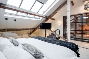 Ett sovrum med ett takfönster, en säng med vita sängkläder, en grå pläd och en tv som är monterad på väggen.