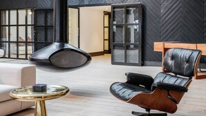 Un interior espacioso con techo alto, vigas de madera a la vista, una gran chimenea negra y una zona de estar moderna con un sillón de cuero y una mesa de vidrio.