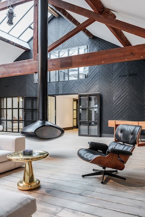 Un interior espacioso con techo alto, vigas de madera a la vista, una gran chimenea negra y una zona de estar moderna con un sillón de cuero y una mesa de vidrio.