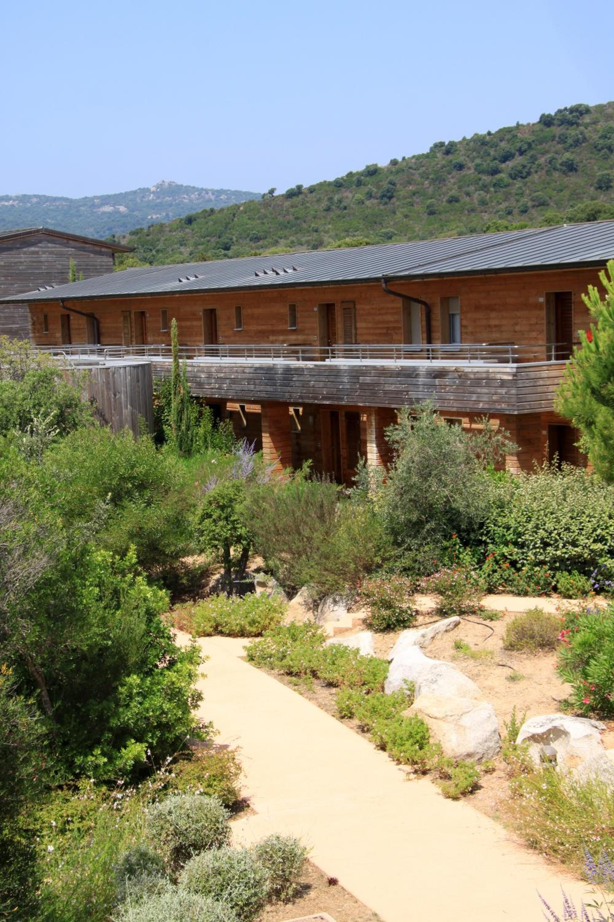 A rustic wooden building with a flat roof, surrounded by a landscaped garden with various shrubs and a gravel path.