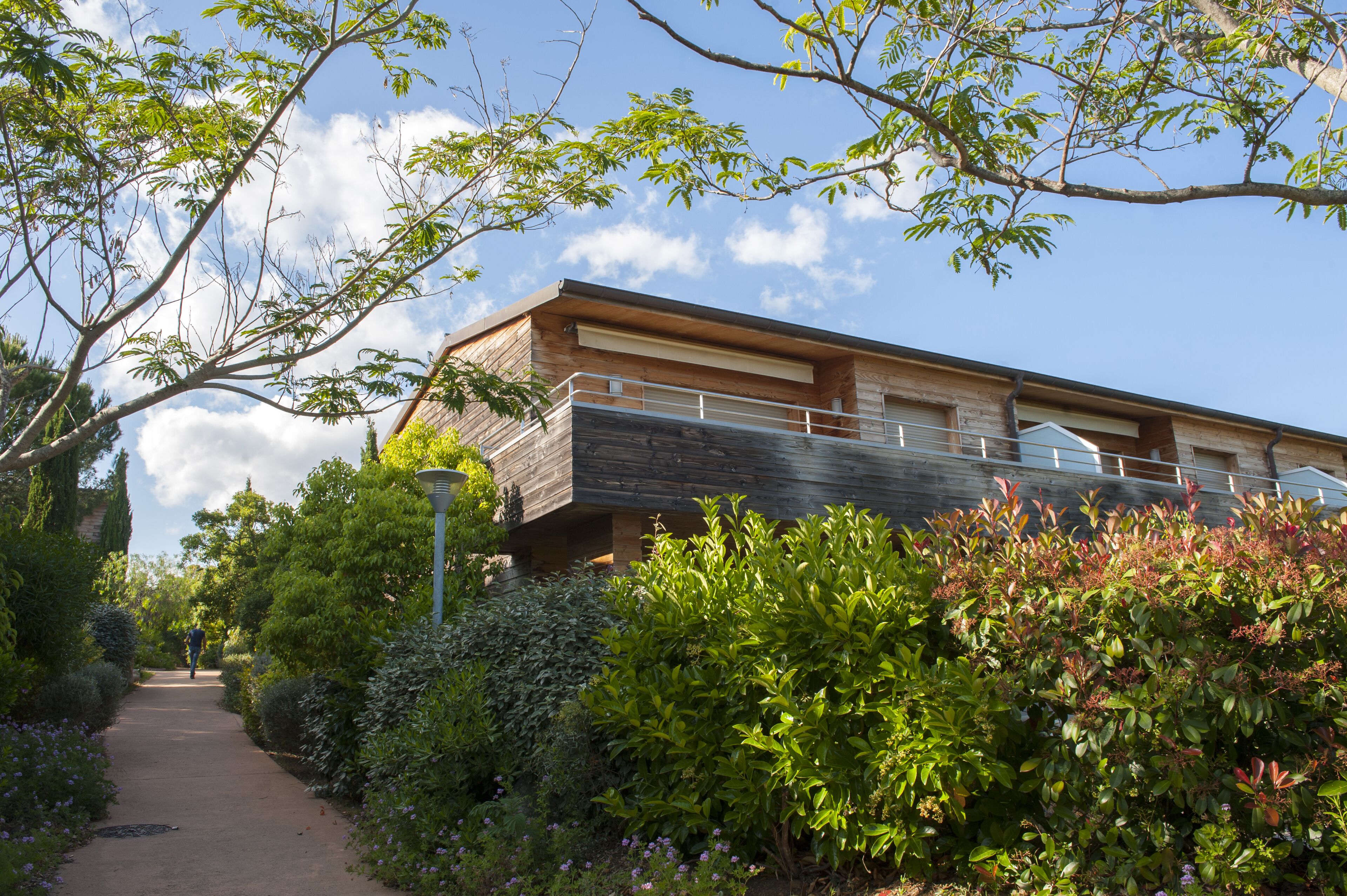 A modern building with a flat roof and a balcony, surrounded by lush greenery and a clear sky.