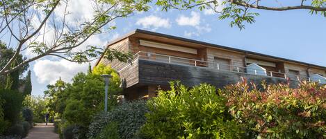 A modern building with a flat roof and a balcony, surrounded by lush greenery and a clear sky.