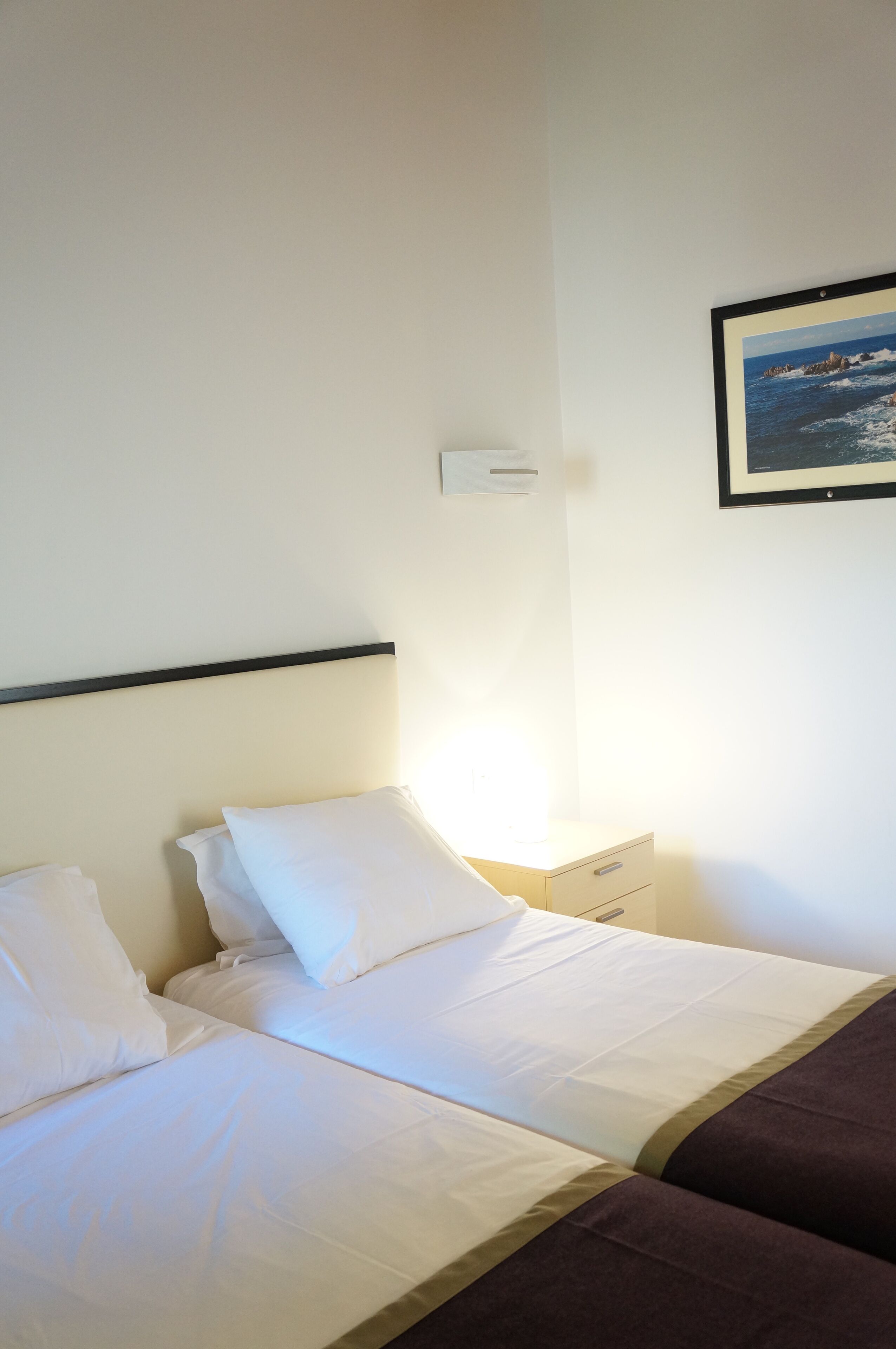 Una habitación de hotel con dos camas, una mesita de noche, una lámpara de pared y un cuadro enmarcado.