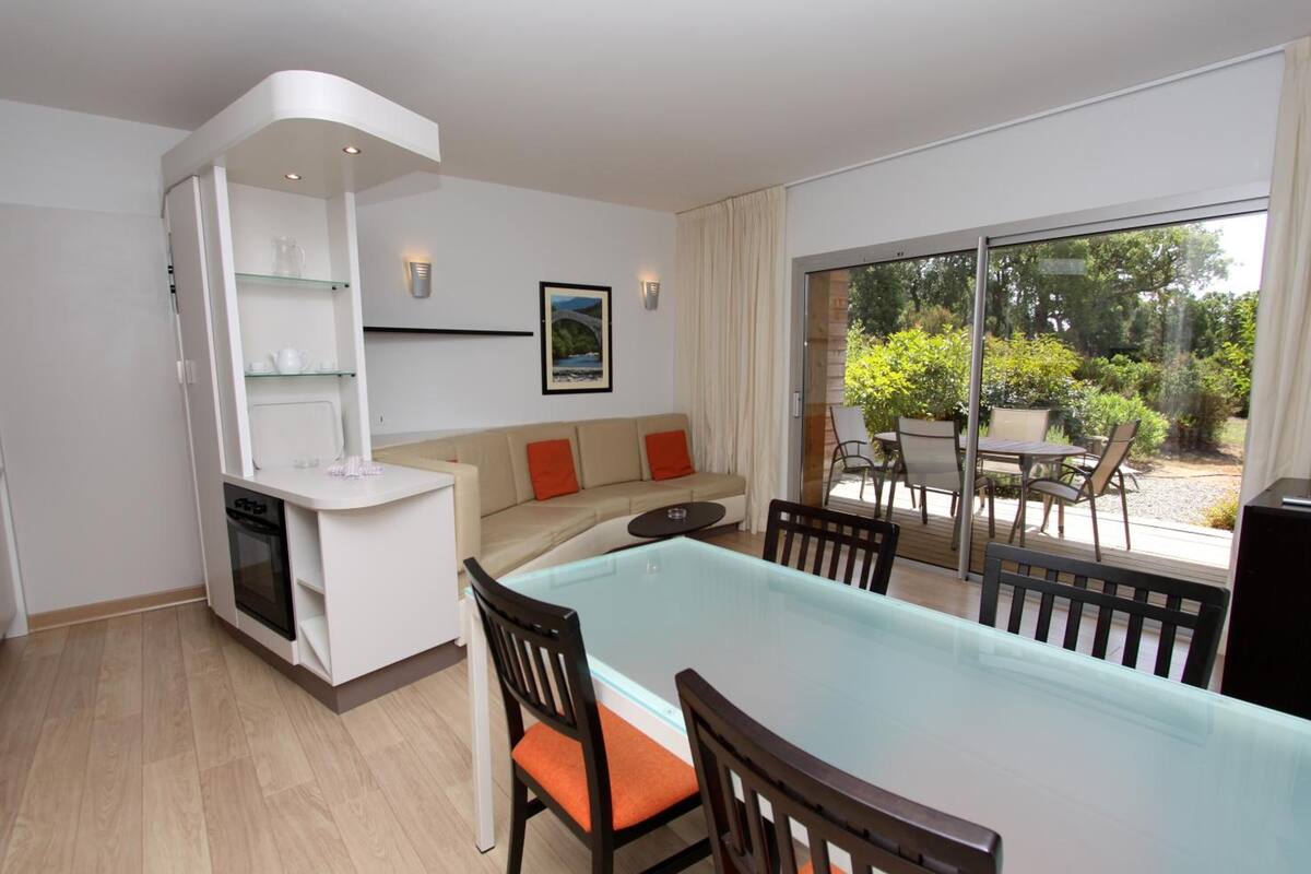 Een ruimtelijk ingerichte leefruimte met kitchenette, een eethoek met glazen tafel en een balkon met tuinmeubilair.