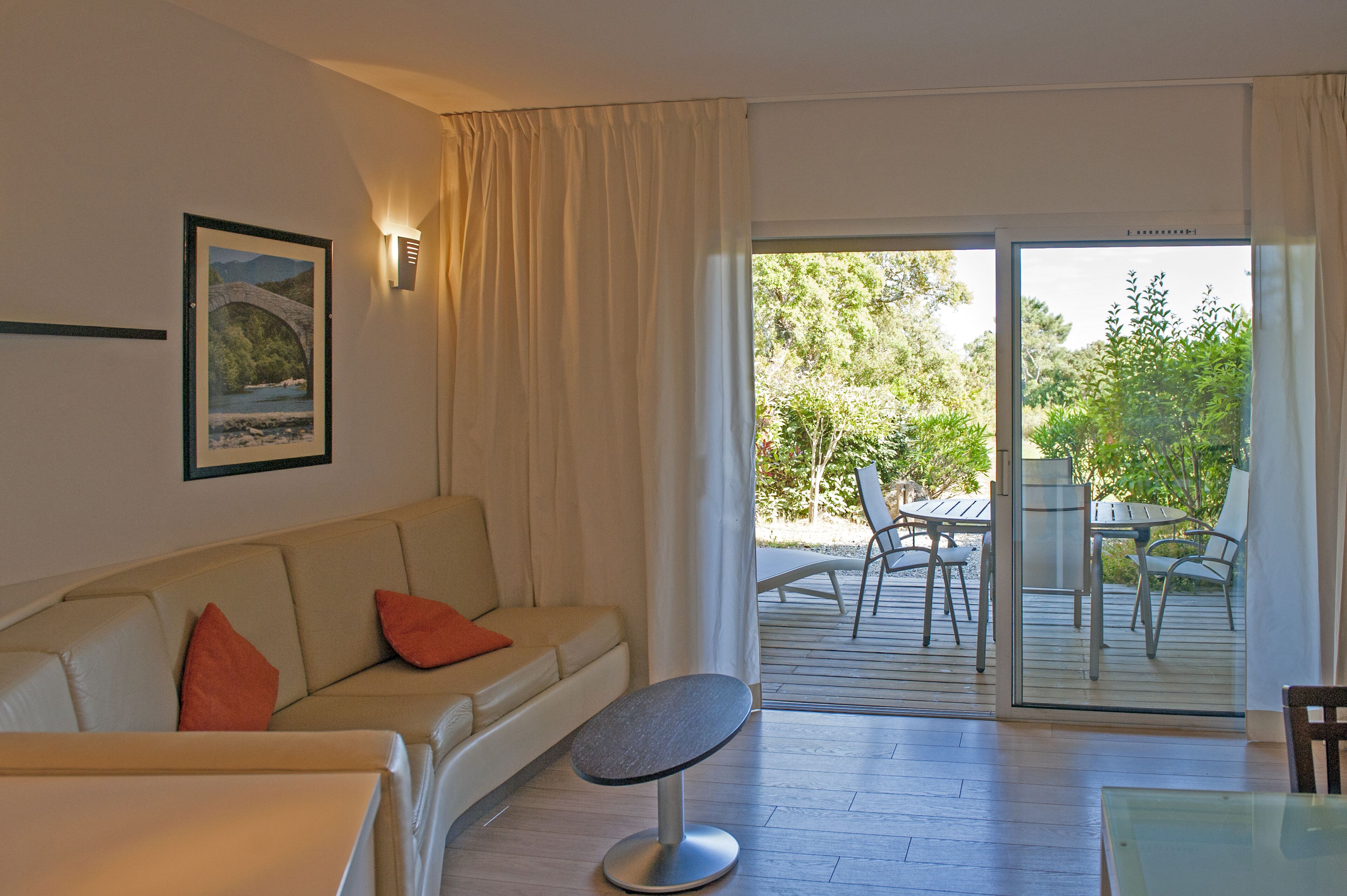 Un salon moderne avec un canapé, une petite table ronde et une table avec un plateau en verre. Une porte-fenêtre coulissante donne sur une terrasse extérieure équipée de chaises et d’une table. Un tableau encadré est accroché au mur.