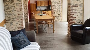 Uma sala de estar com parede de pedra, área de jantar com armários de madeira e cozinha visível por um arco.