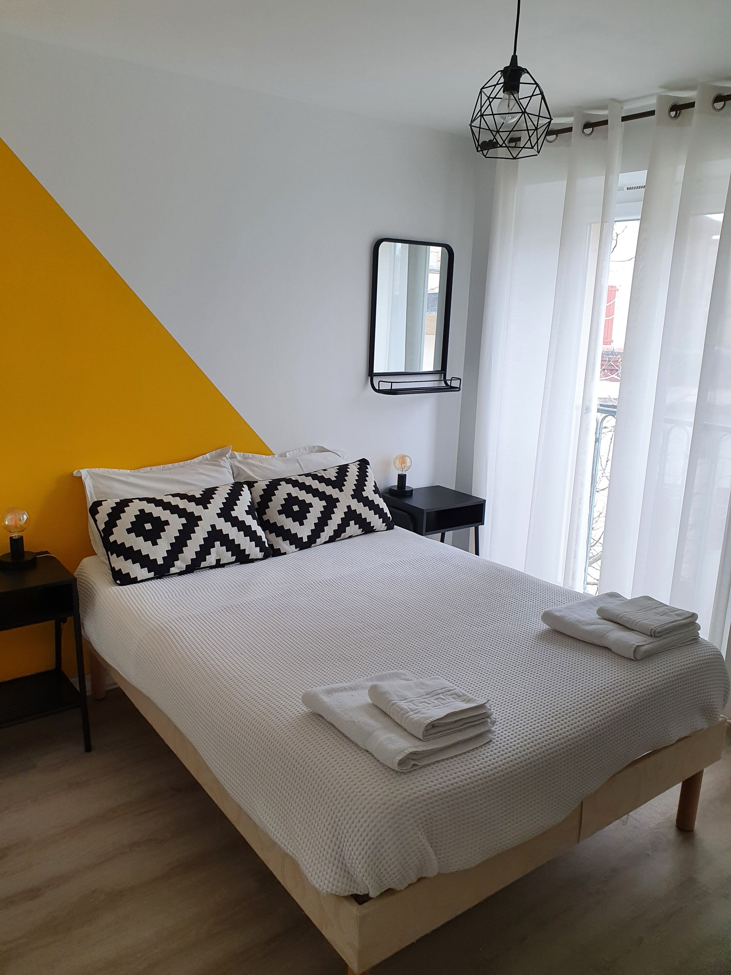 A neatly made bed with a geometric-patterned pillow, a bedside table with a lamp, and a mirror on the wall.