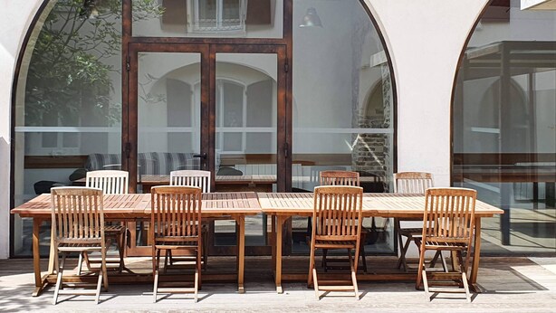 Uma mesa de madeira com seis cadeiras posicionadas em frente a uma parede com amplas janelas em arco.