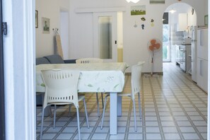 A dining area with a table and chairs, a kitchen in the background, and a tiled floor. - M107 - Marcelli, three-room apartment with garden 200m from the sea (Marcelli)