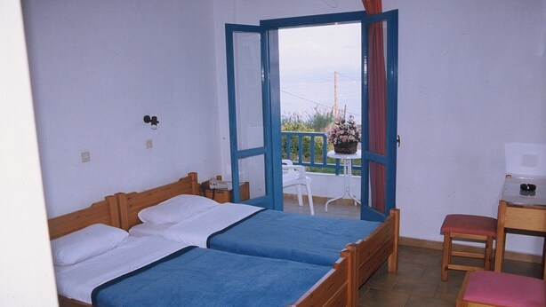 A room with two wooden beds, a small table with a chair, and a view of a balcony with a potted plant. - Friday Hotel (Hersonissos)