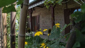 Um edifício rústico com fachada de tijolos e vigas de madeira, rodeado por bambu e flores amarelas.