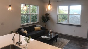 A modern living room with a black sofa, a coffee table, and a kitchen area with a sink. There are large windows providing views of trees and a building. - 750 Square Feet, 1 Bedroom - Walk to Everything! Great for longer term stays. (Austin)