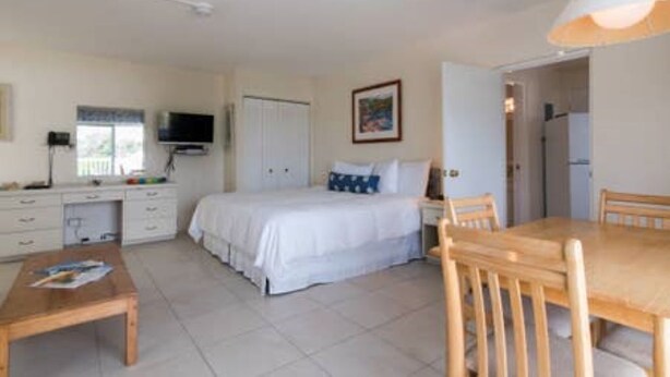 A compact bedroom with a bed, bedside table, and a desk with a chair. There is a window with blinds and a door leading to another room. - Clairfont, Jobson's Cove / Warwick Long Bay/ Horseshoe Beach -  APARTMENT 1 (Warwick)