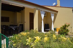 A covered outdoor seating area with a table and chairs, a small garden with yellow flowers, and a building with a satellite dish. - Bilocale Riccardo (Muravera)