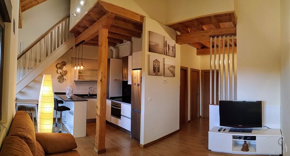 Uma cozinha moderna com vigas de madeira, uma sala de jantar com um sofá e uma TV sobre um suporte.