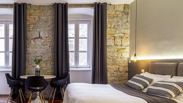 A hotel room with a stone wall, a bed with striped pillows, a round table with chairs, and a vase with a plant. - Romantic Studio in the heart of Trieste ★Free Wifi (Trieste)