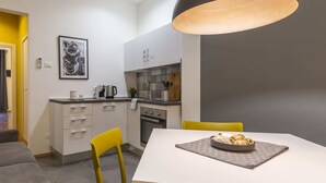 Una cocina compacta con área de comedor, que incluye una mesa blanca con mantel individual estampado, una silla amarilla y una lámpara colgante negra.