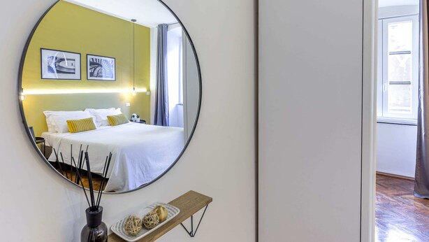 Habitación de hotel con un espejo grande y redondo, una cama con ropa blanca, un estante con objetos decorativos y cuadros en la pared.