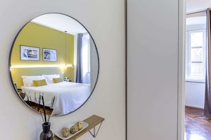 A hotel room with a large round mirror, a bed with white bedding, a small shelf with decorative items, and framed pictures on the wall.