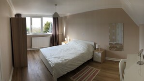 A bedroom with a bed, bedside table, lamp, and a window with curtains. - Little Mead (Tenby)