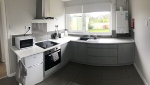 A modern kitchen with a microwave, toaster, and oven, a window with a view of the garden, and a sink. - Little Mead (Tenby)