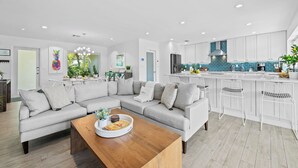 A modern living room with a sectional sofa, a wooden coffee table, and a kitchen with white cabinets and a blue backsplash.