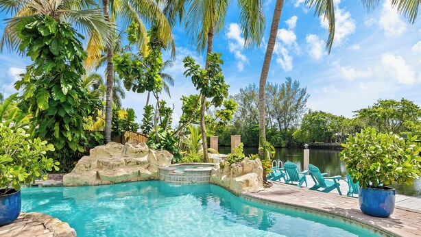 A clear swimming pool with a stone edge, surrounded by palm trees and a wooden deck with chairs.