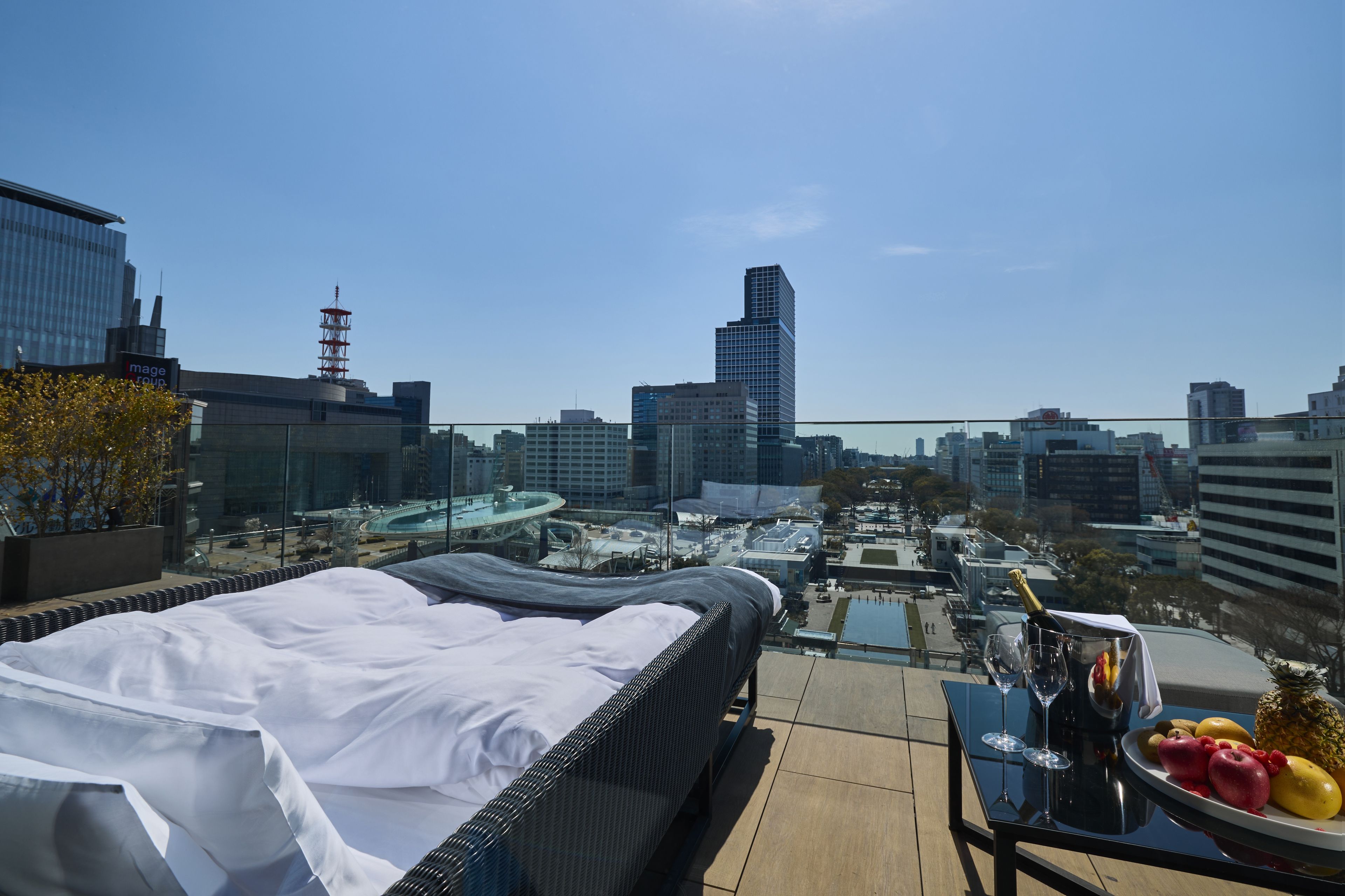 A rooftop terrace with a bed, a table with a bottle and glasses, and a view of the cityscape.