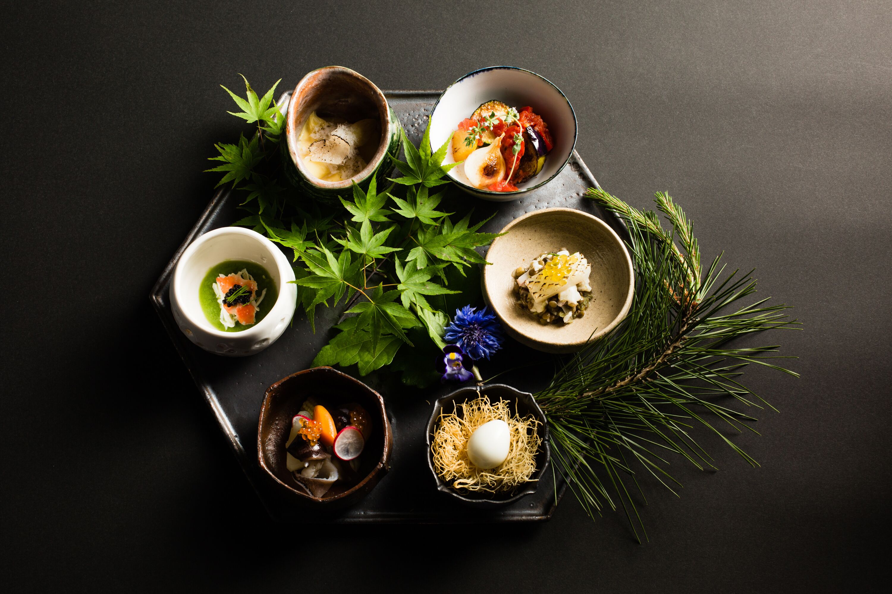 A tray with five bowls of food, each containing different dishes, accompanied by green leaves and pine branches.