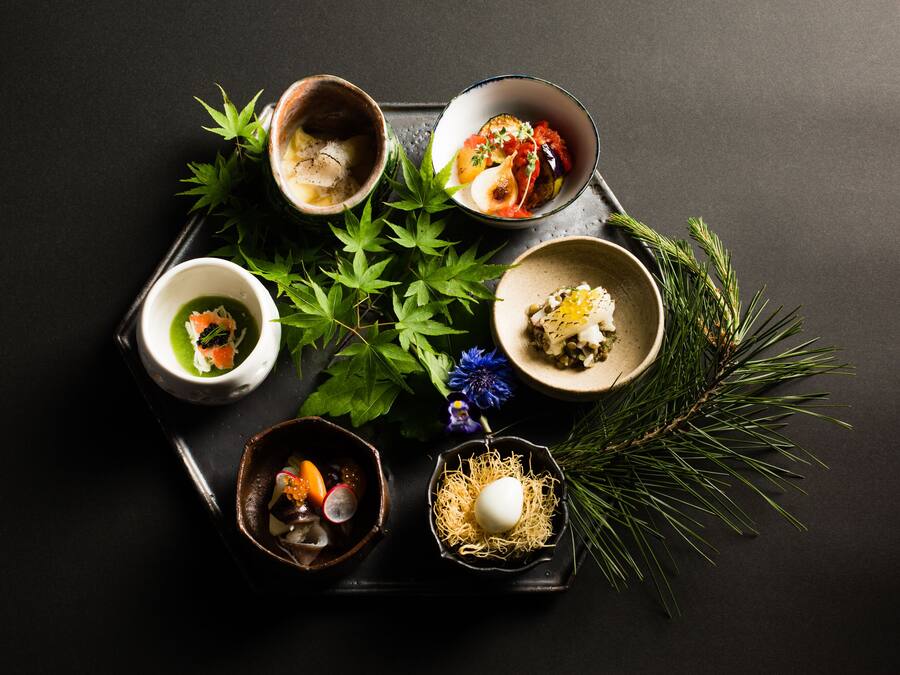A tray with five bowls of food, each containing different dishes, accompanied by green leaves and pine branches.