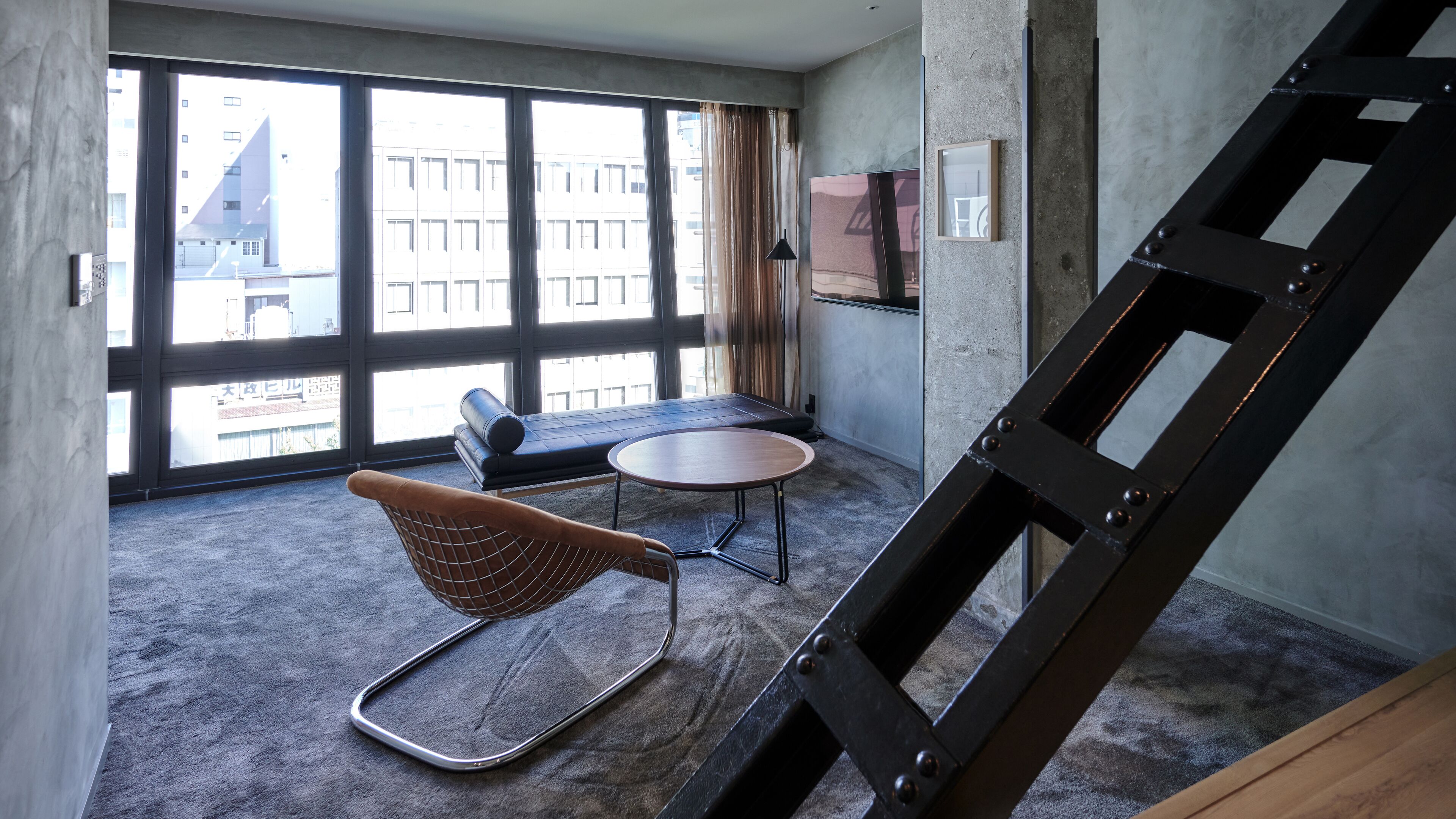 Ein modernes Zimmer mit großen Fenstern, einem runden Tisch, einem gepolsterten Sessel und einer Metallleiter.