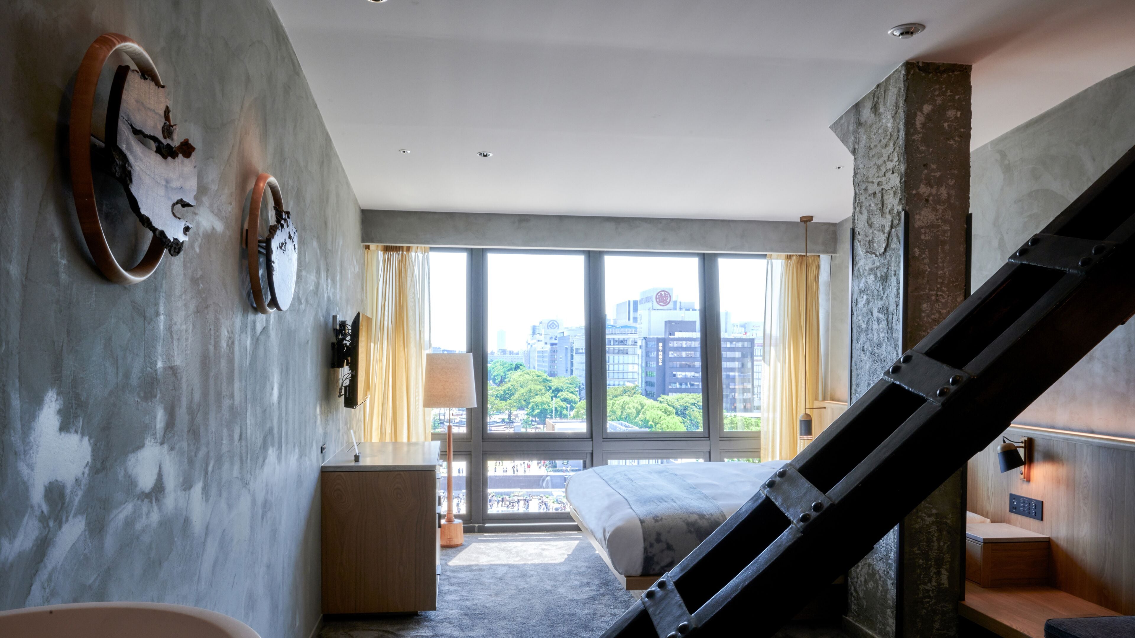 A modern hotel room with a large window, a bed, a desk, and a staircase.