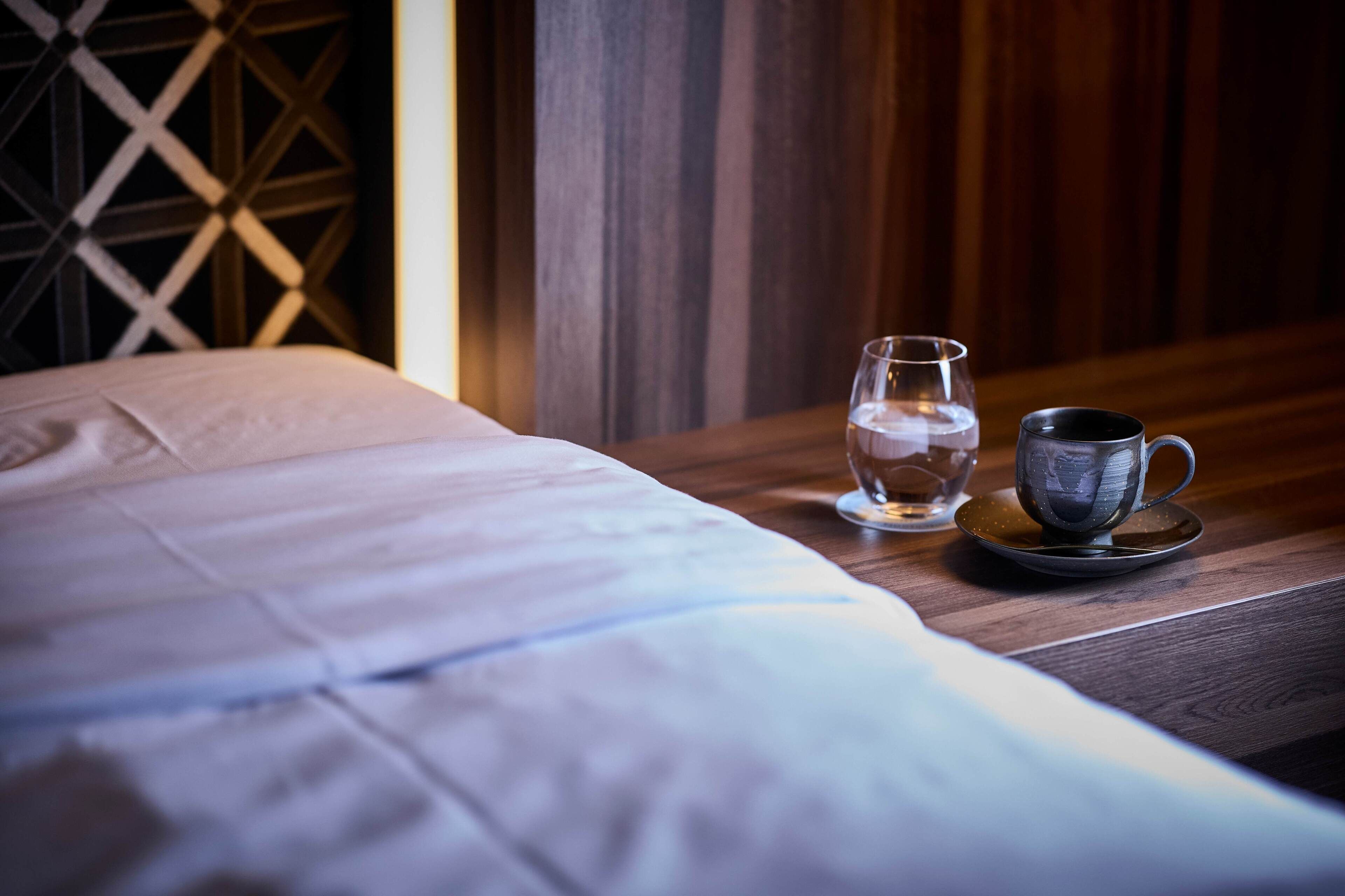 Ein ordentlich bezogenes Bett mit weißer Decke und ein Nachttisch daneben. Auf dem Nachttisch steht ein klares Wasserglas und eine schwarze Tasse auf einem Untersetzer.