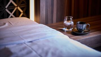 Ein ordentlich bezogenes Bett mit weißer Decke und ein Nachttisch daneben. Auf dem Nachttisch steht ein klares Wasserglas und eine schwarze Tasse auf einem Untersetzer.