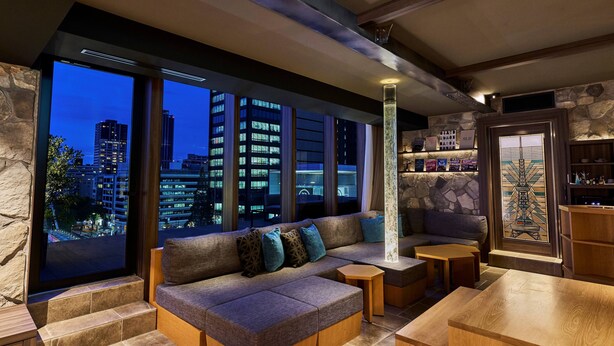 A modern living room with a large window offering a city view, a cozy sectional sofa, and a small wooden table. - The Tower Hotel Nagoya (Nagoya)