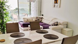 A modern living room with a white sofa, a dining table with chairs, and a large window with curtains. - Fine apartment by the sea, beach, nice and quiet yet close to Center (Villajoyosa)