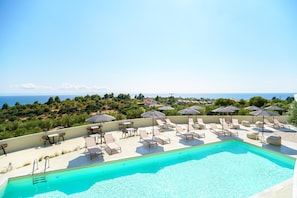 Piscina en la azotea con sillones y sombrillas, rodeada de vegetación y con vistas a un paisaje costero.