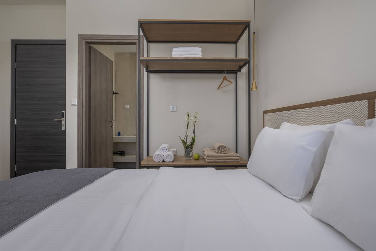 Une chambre d’hôtel avec un lit, une table de chevet en bois sur laquelle se trouvent des serviettes et un vase de fleurs, une lampe suspendue et une armoire.