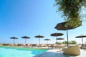 Zona junto a la piscina con sillones de descanso y sombrillas, cielo despejado y una piscina.