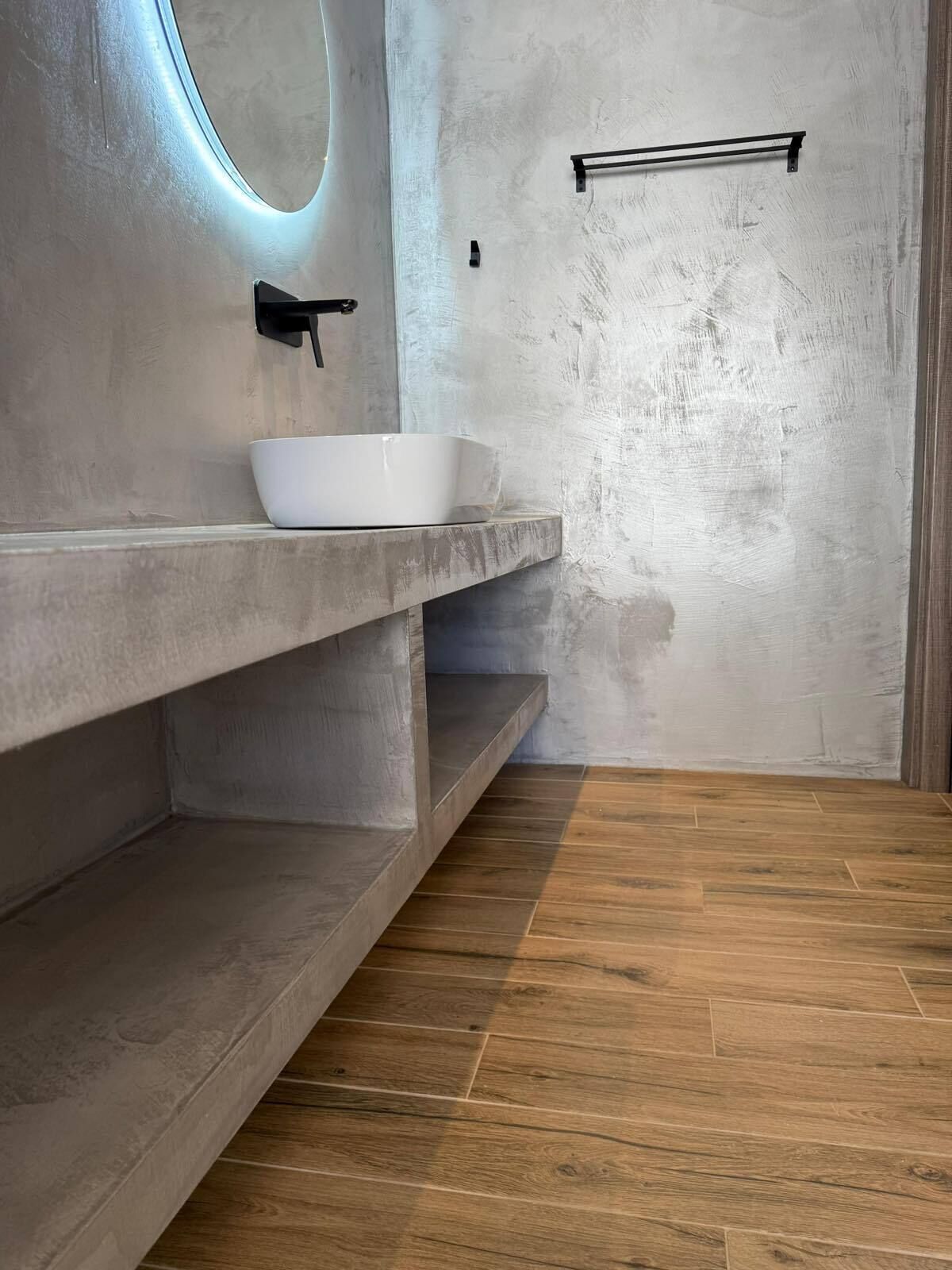 Ein modernes Badezimmer mit einem Betontischwaschbecken, einem runden Spiegel und einem Holzboden.
