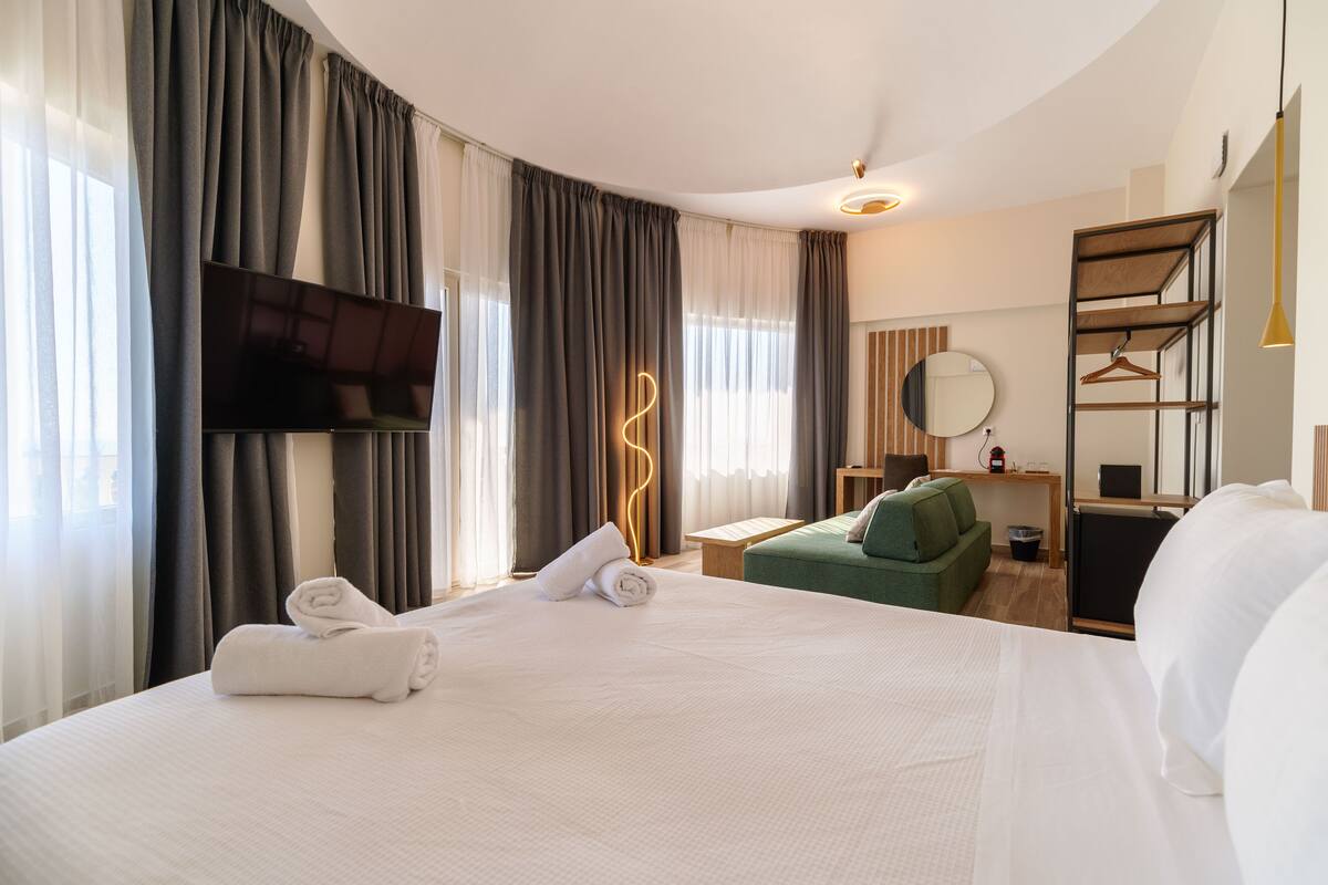 Une chambre d’hôtel moderne avec un grand lit, un téléviseur à écran plat, un canapé vert, une petite table et un miroir.