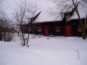 A snow-covered wooden house with red doors and windows, surrounded by bare trees and a snowy landscape.