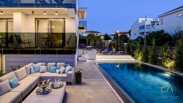 A modern outdoor pool area with a sofa and lounge chairs, a glass balustrade, and a view of the surrounding neighborhood.