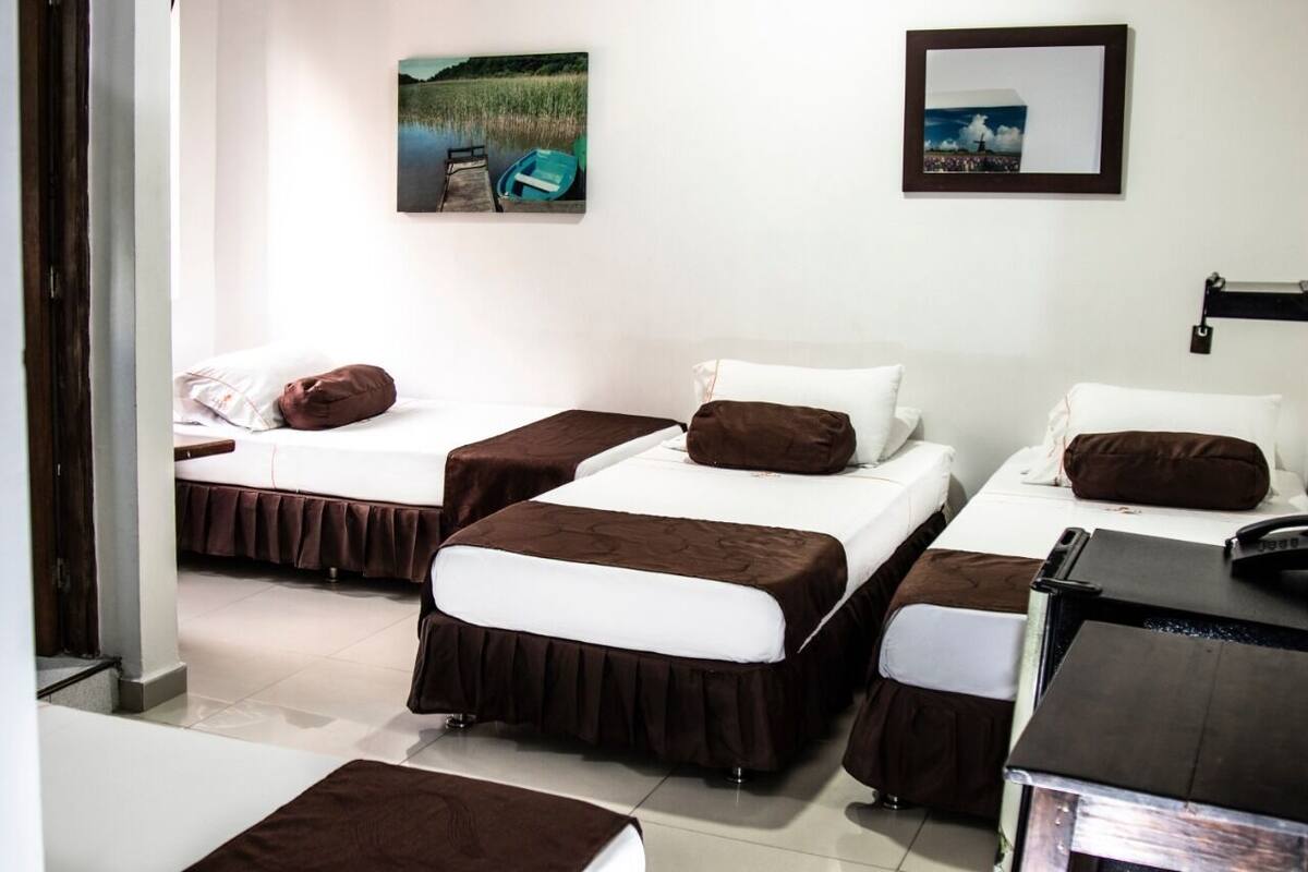 Habitación de hotel con tres camas, cada una con cubrecama marrón y almohadas blancas. Hay dos cuadros en la pared, uno que muestra un barco en el agua y el otro un paisaje urbano.