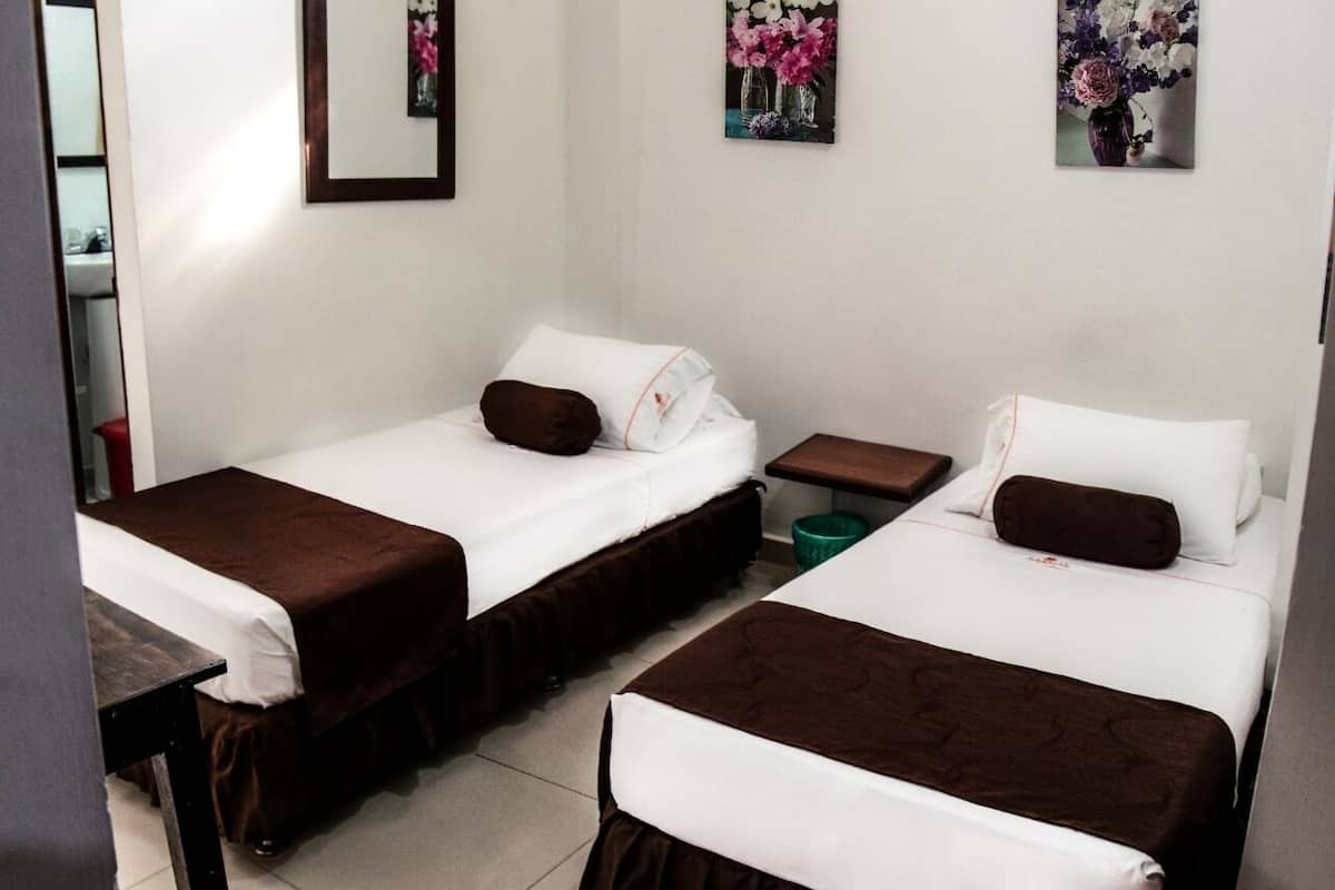 Habitación de hotel con dos camas individuales, cada una con cabecero marrón y ropa de cama blanca.  Entre las camas hay una mesita de noche de madera, y un cubo de basura verde a un lado.