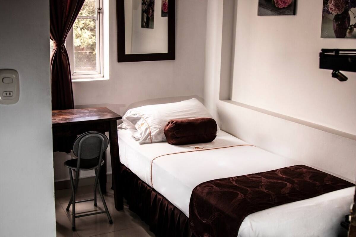 Una habitación pequeña con cama individual, escritorio y silla, ventana con cortinas y estante en la pared.