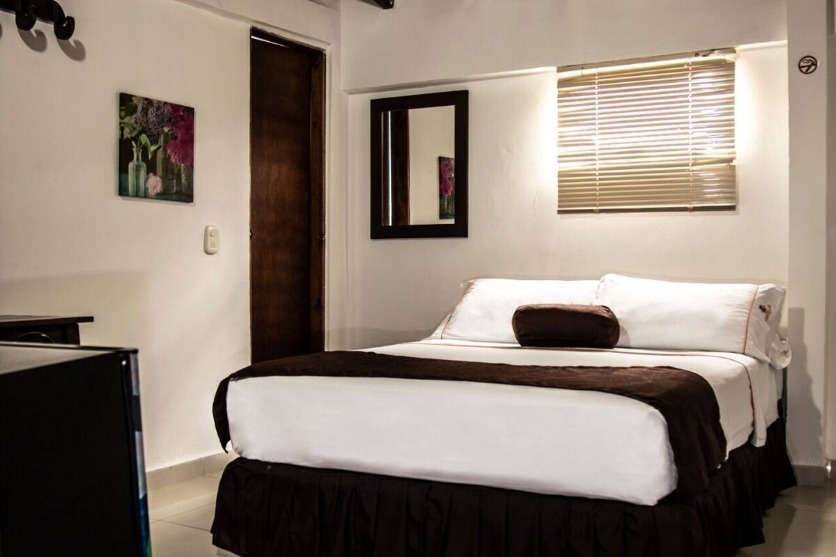 Un dormitorio con cama, espejo, ventana con persianas, un cuadro en la pared y una puerta.