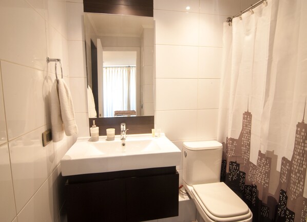 A bathroom with a white sink, mirror, and toilet. There is a towel rack on the wall and a window with curtains. - Views Cordillera (Santiago)