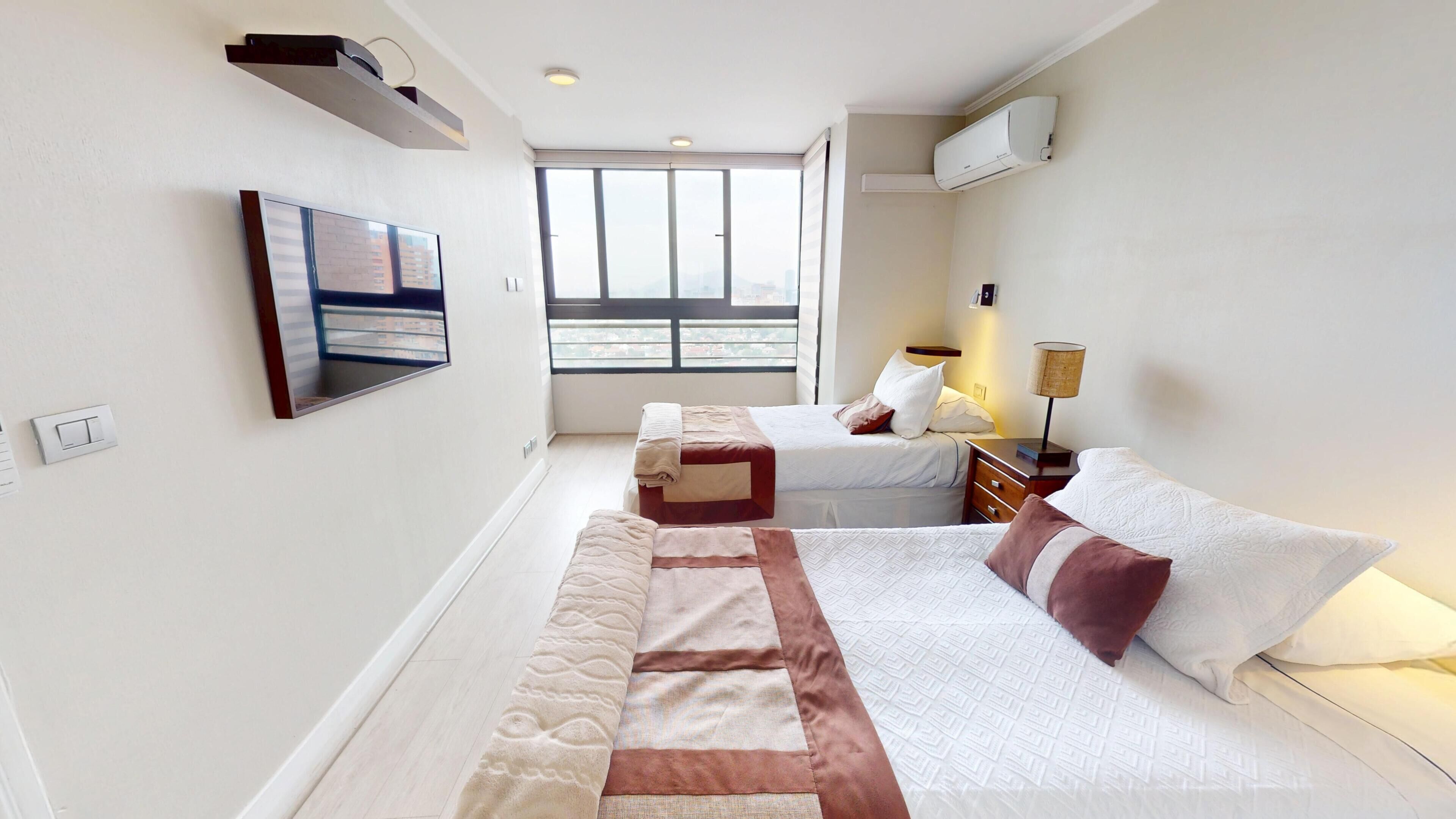 Habitación de hotel con cama, mesita de noche con lámpara, televisión montada en la pared y ventana con vista a la ciudad.