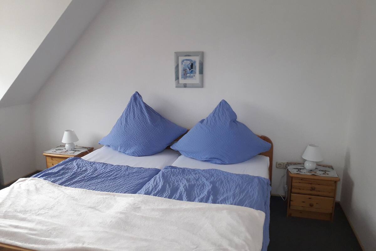 A bedroom with a sloped ceiling, a bed with blue and white bedding, two bedside tables with lamps, and a framed picture on the wall.